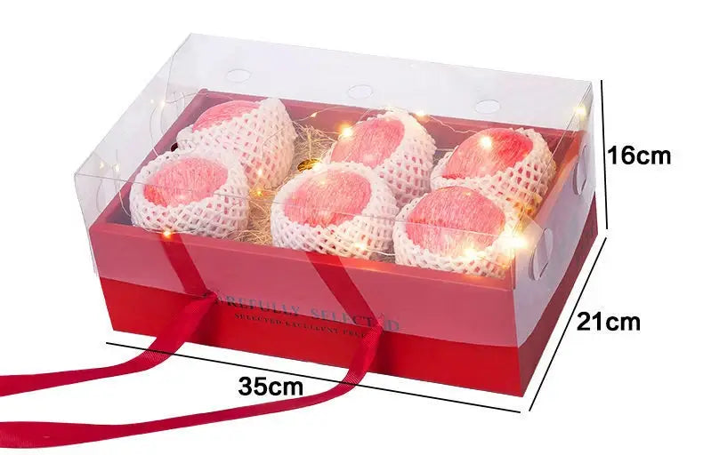 red bow knot handheld flower box in red with a clear plastic lid containing six pink fruits wrapped in white mesh netting and decorated with string lights.