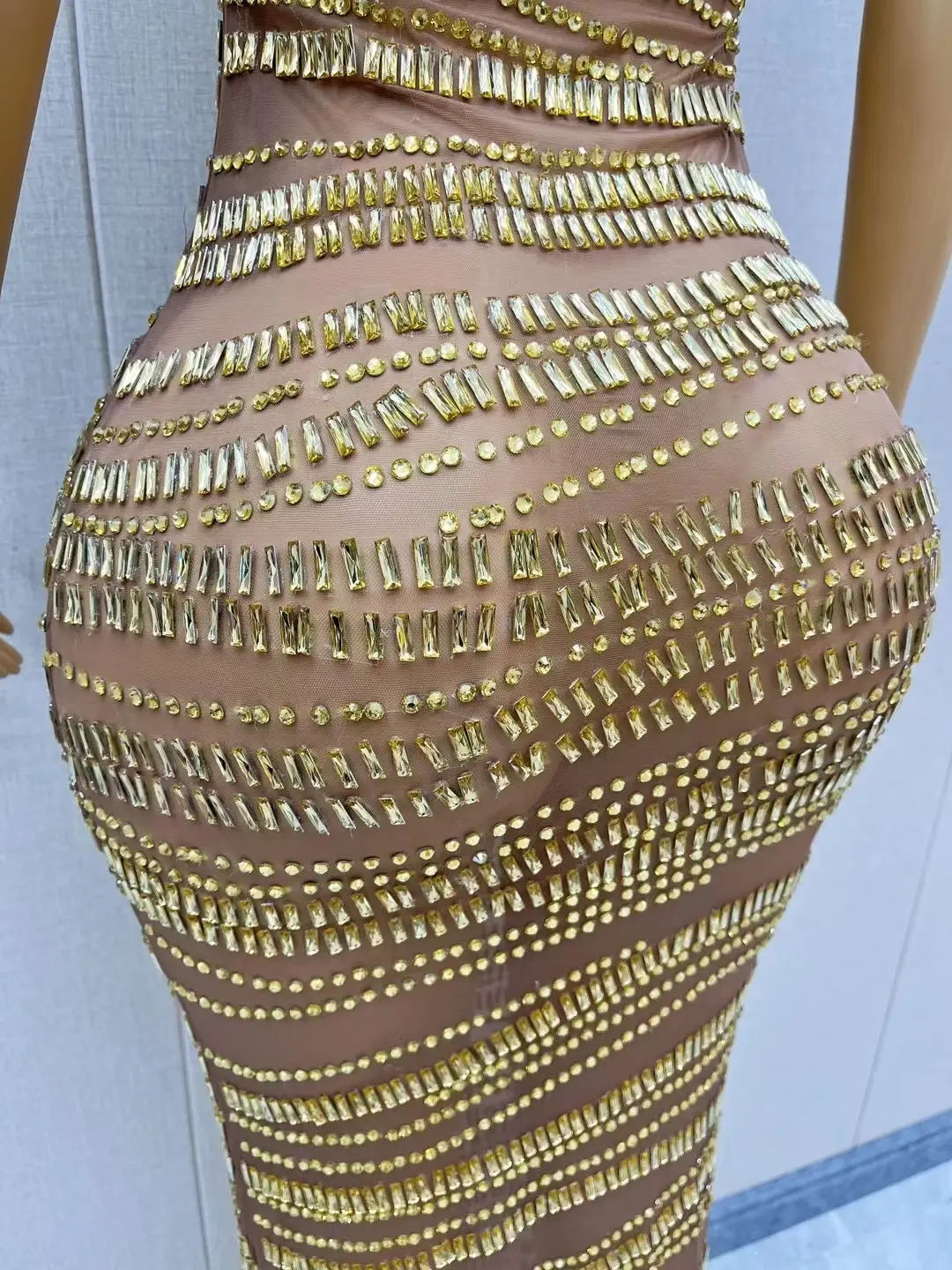 A luxury shiny rhinestone dress featuring a sheer gold fabric with form-fitting design and intricate rows of gold-toned rhinestones and smaller circular embellishments.