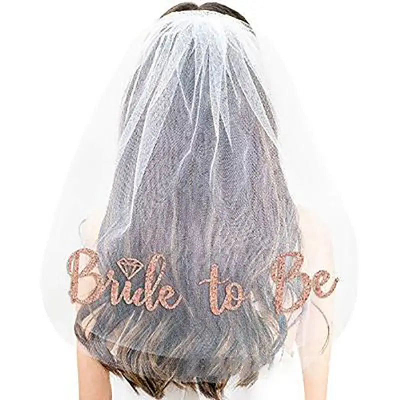 A delicate sheer white bridal veil popular made of tulle features rose gold glitter lettering reading bride to be and a diamond-shaped embellishment.