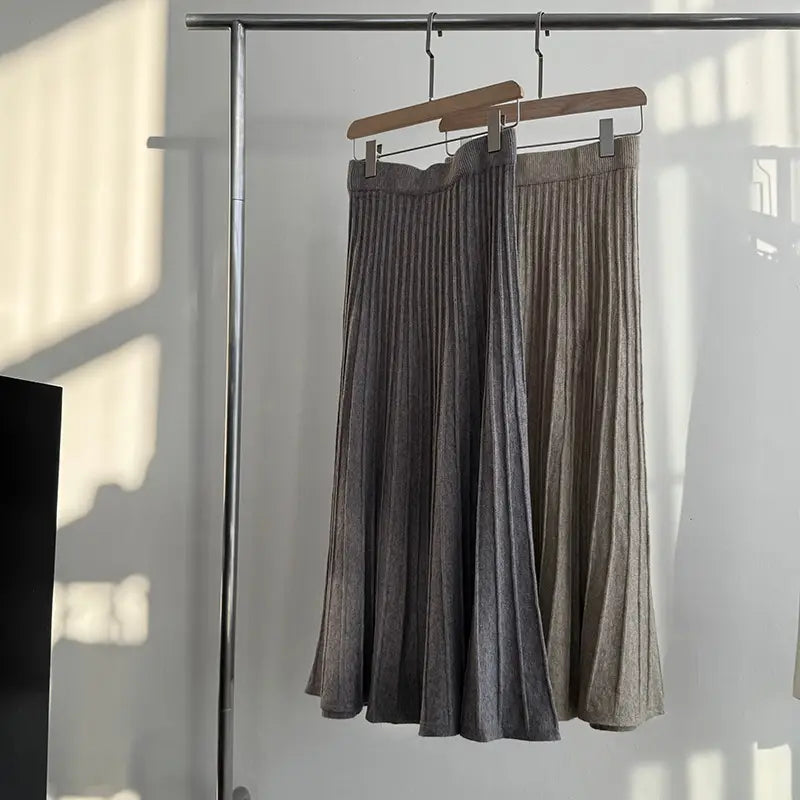 two korean elastic waist knitted pleated midi skirts in muted gray and soft beige hang on wooden hangers from a metal rack.