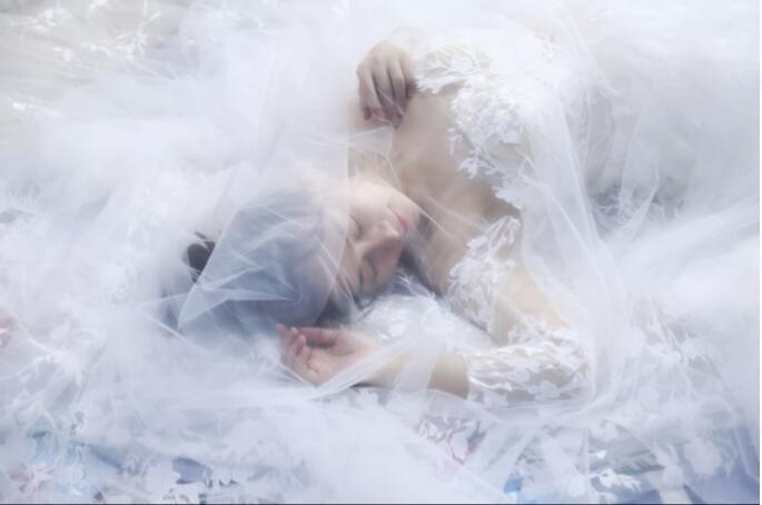 a woman lies nestled in a sea of sheer translucent white lace fabric showcasing a bridal wedding veil with a widened tail made of lace