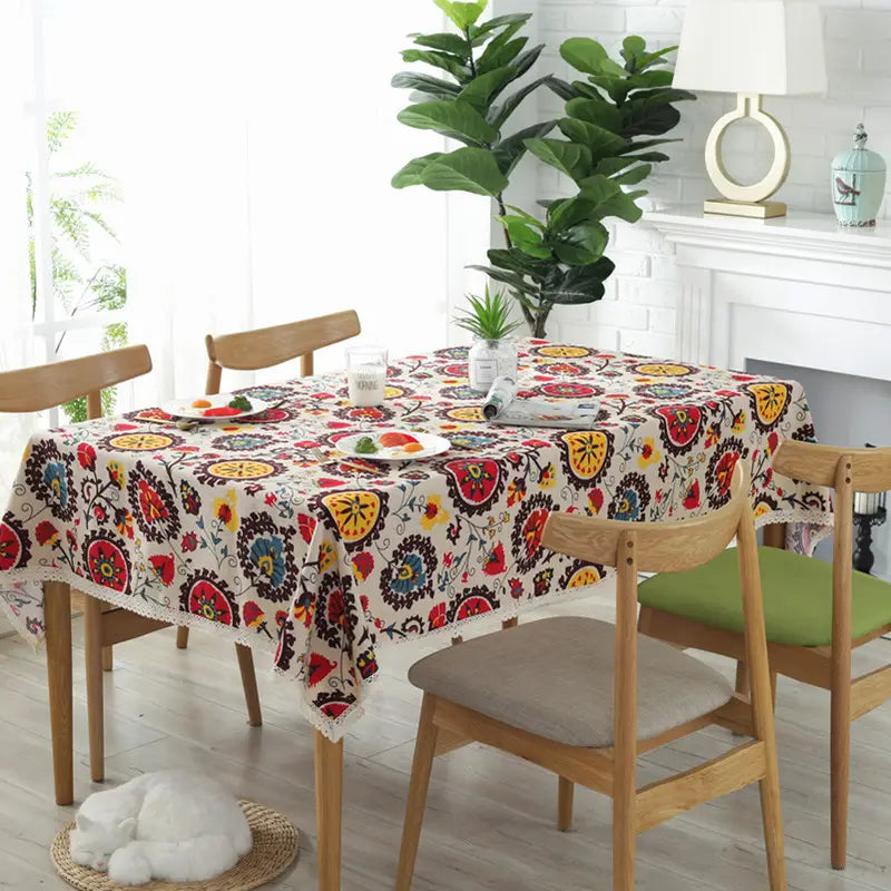 A vibrant linen table cloth with colorful floral and geometric sunflower cotton designs in reds yellows blues and browns on a cream background featuring delicate lace trim along the edges.