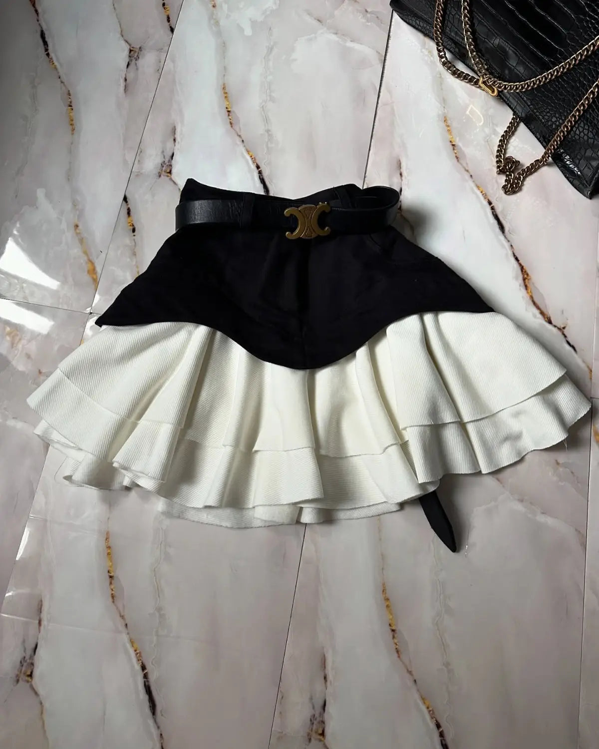black and white two-tone pleated mini skirt with a black leather belt featuring a gold-toned c-shaped buckle resembling a high waist tiered skirt design.