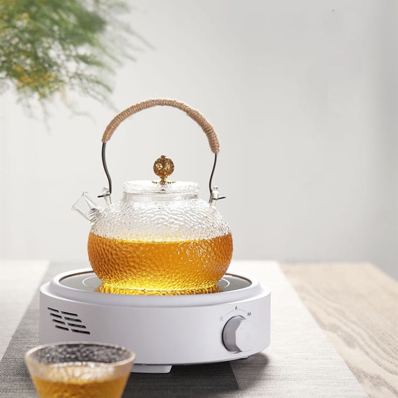 white ceramic electric earthenware oven featuring a clear textured glass teapot with a woven handle and golden lid knob on a white electric heating base.
