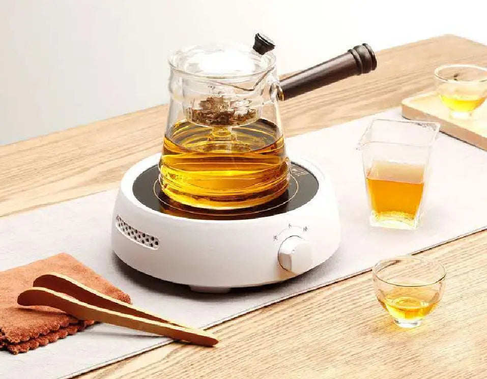 clear glass teapot with dark wooden handle and black lid knob filled with golden tea on white ceramic electric earthenware oven heating base