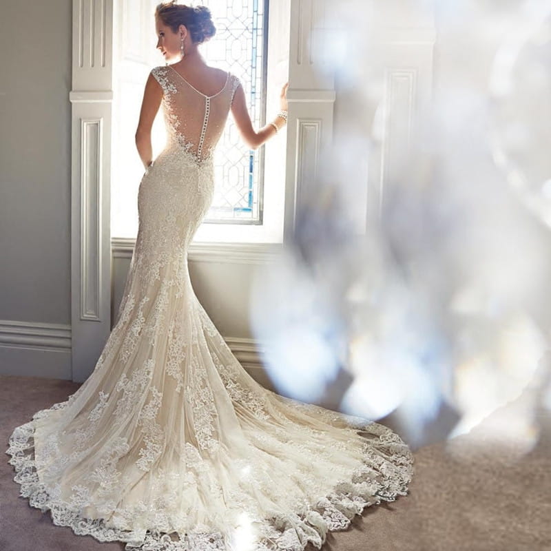 A woman wears an ivory lace sexy halter brigade shimmering mermaid wedding dress featuring a beaded back and flowing train shown from the back side.
