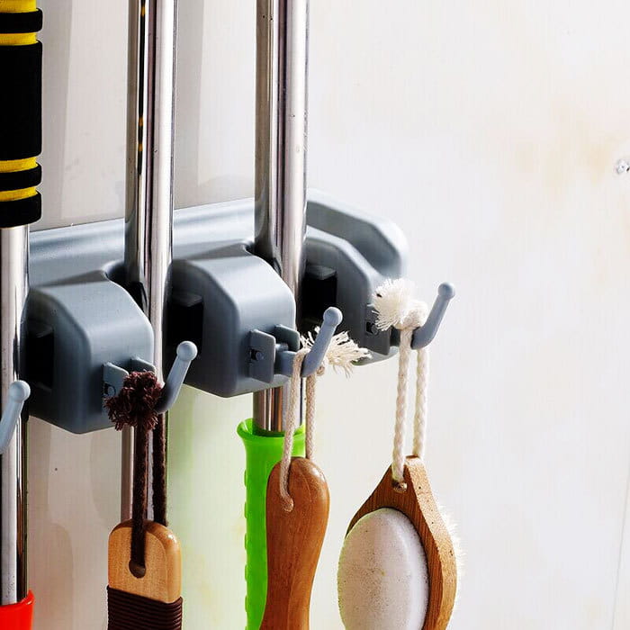 Gray plastic mop hook wall hanging organizer rack with multiple slots and hooks for handles mounted on a wall