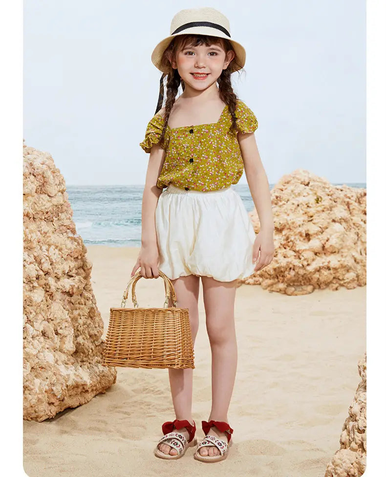 Woven wicker handbag with natural tan handles and a rectangular body displayed alongside big children’s soft-soled embroidered floral sandals.