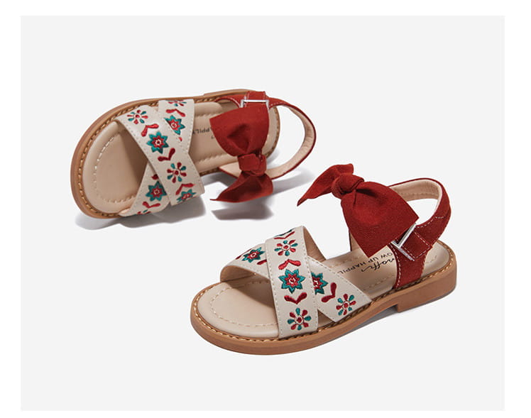 cream-colored big children’s soft-soled sandals with red and teal floral embroidery and a matching red satin bow on the ankle strap designed for baby children’s shoes.