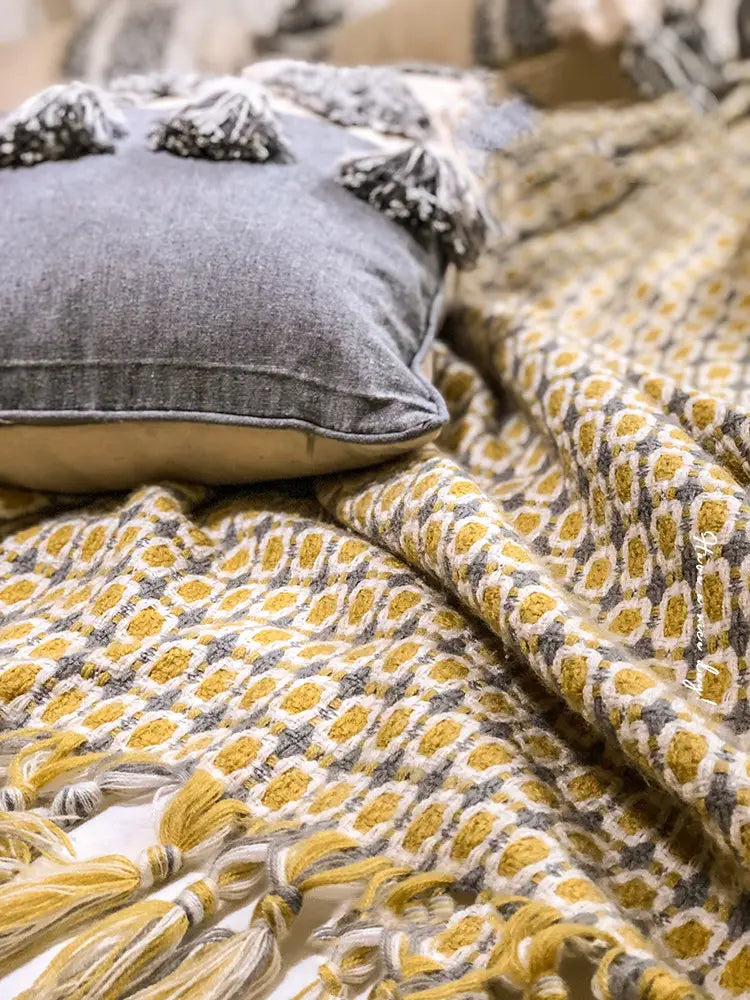 gray cushion with black tassels resting on a mustard geometric knitted sofa blanket cover with tassels featuring a yellow and gray patterned blanket cover blanket