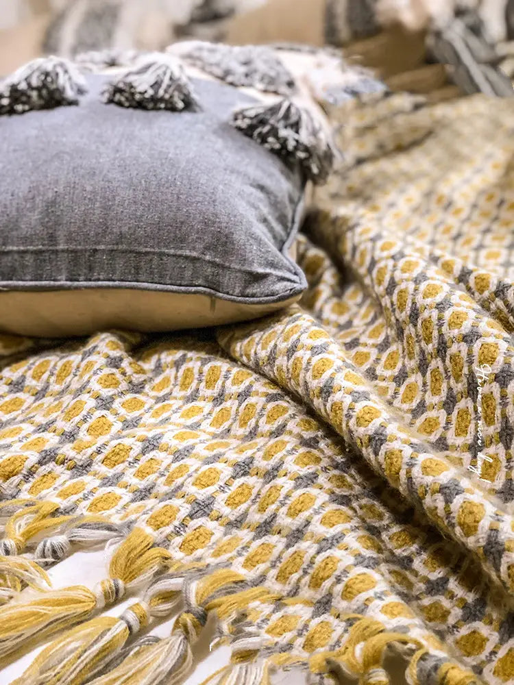gray denim pillow with black tassels resting on a mustard geometric knitted sofa blanket cover with tassels and a yellow and gray patterned blanket cover blanket