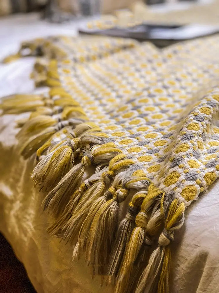 a cozy mustard-yellow and cream-colored cover blanket knitted with a geometric pattern and finished with braided tassels along the edge, ideal as a sofa blanket cover.