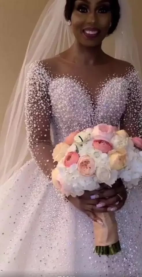 A sparkling lavender and white beaded ball gown wedding dress with long sleeves featuring a fitted bodice adorned with intricate pearl-like embellishments.