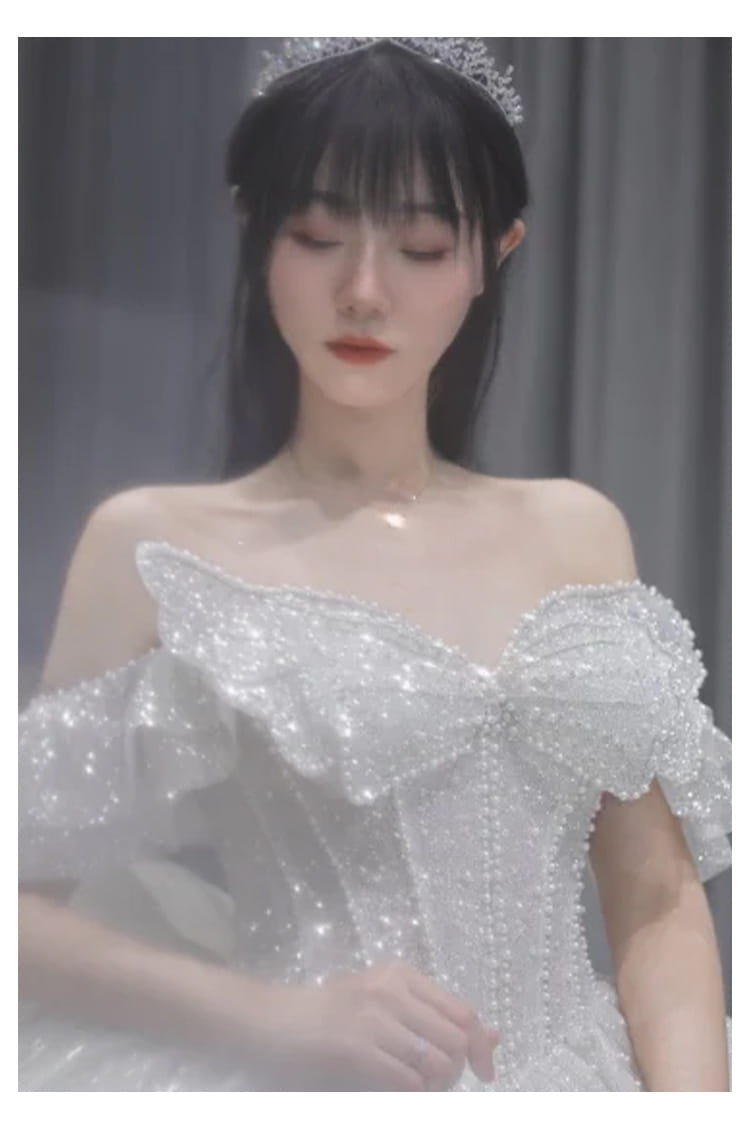 A sparkling white off-the-shoulder wedding dress with intricate beading and a large bow at the bodice designed as a long trailing heavy industry gown.