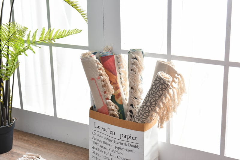 a white paper storage bin with a brown cardboard lid labeled le sac en papier holding rolled-up scandinavian style linen cotton rugs with tassels in various colors and patterns
