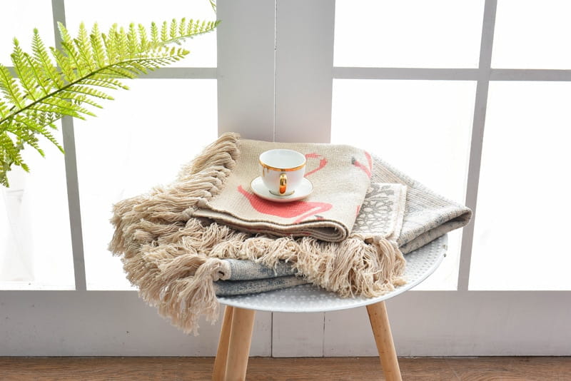 beige throw blanket with fringed edges and subtle pink flamingo design layered over a gray knit piece on a round wooden table showcasing scandinavian style linen cotton rug with tassels