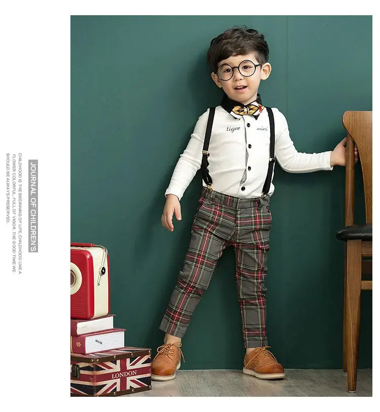 A young boy wearing a men’s plaid suspenders treasure suit tide suit featuring gray and red plaid trousers, white long-sleeved shirt, black suspenders, and a patterned bow tie.