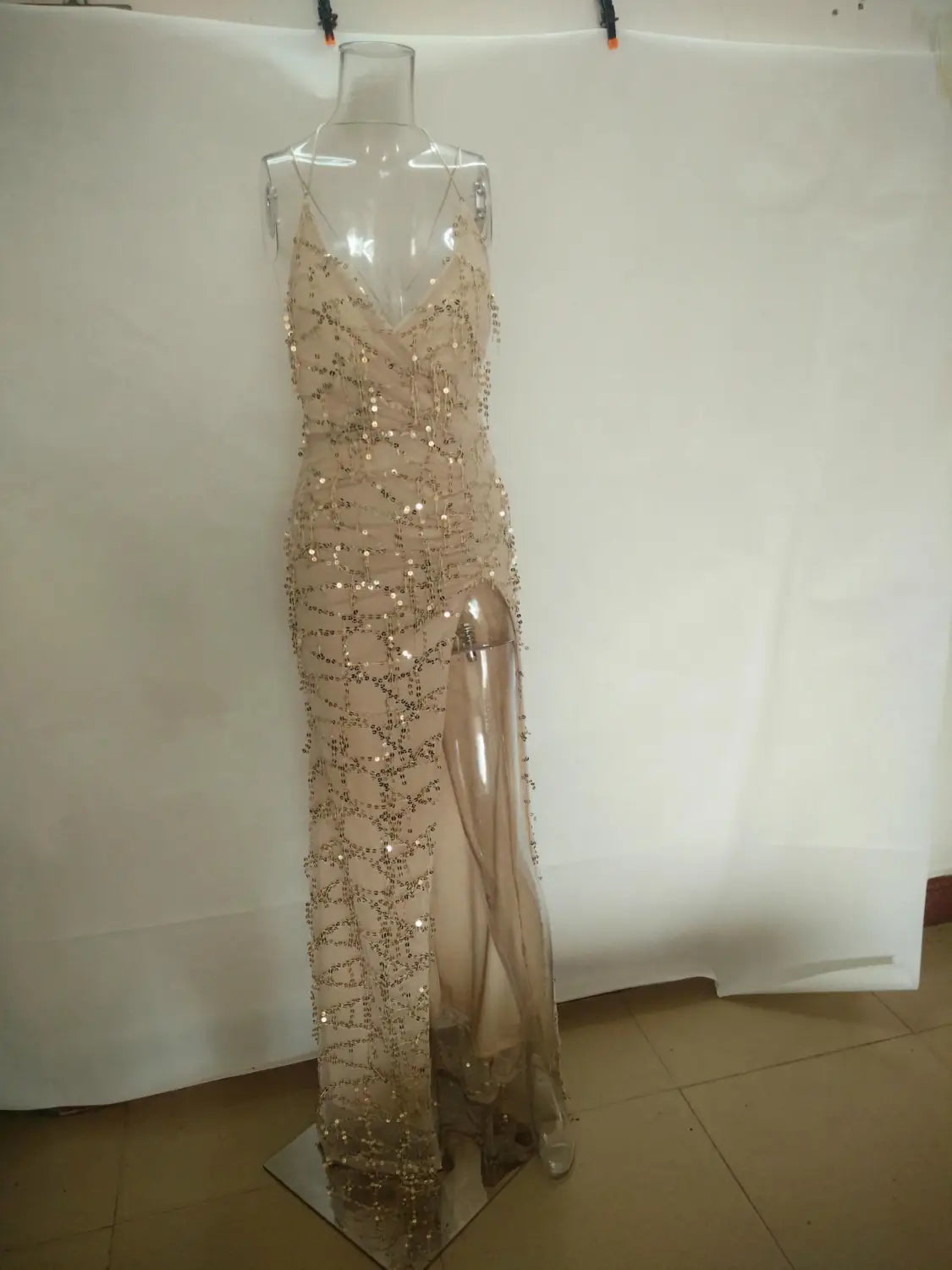 A shimmering champagne-colored gold v neck halter strap sequin dress with intricate beading and a high slit displayed on a clear mannequin.