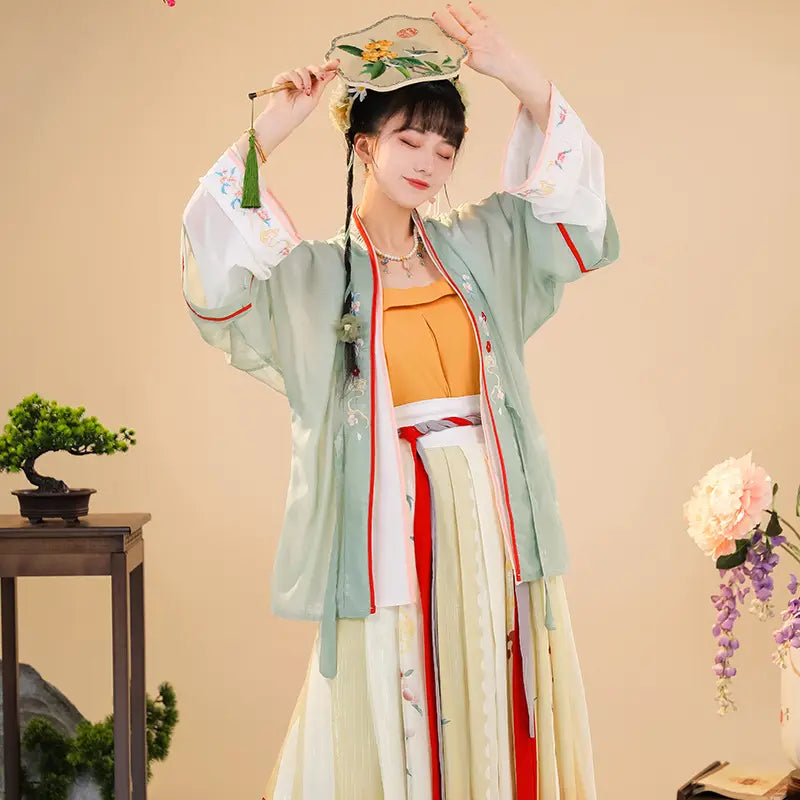 A woman wears a light green embroidered hanfu jacket with red and white trim over an orange top paired with a cream-colored pleated skirt showcasing han chinese clothing summer improvement.