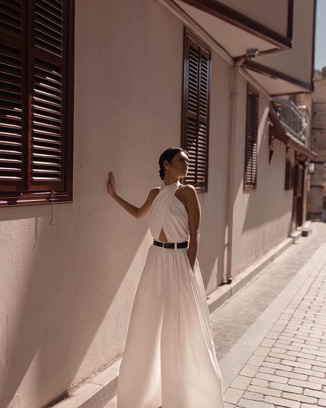 a flowing white sexy sleeveless halter backless jumpsuit with a cutout detail at the back and cinched at the waist by a black belt