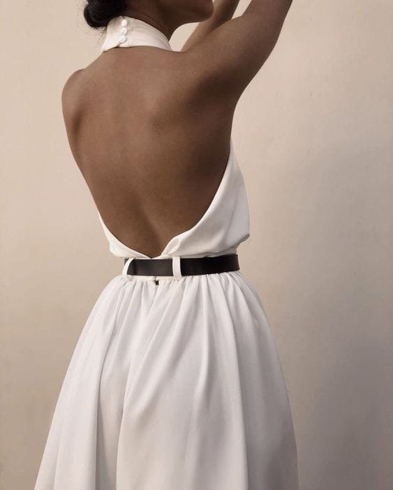 a white sexy sleeveless halter backless jumpsuit with pearl embellishments at the neckline and cinched by a black belt showcasing the back design