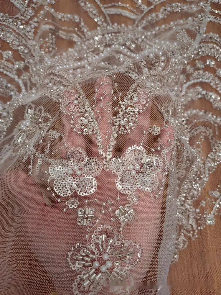 A delicate sheer white lace fabric with intricate silver wave style sequin beadwork forming floral patterns for a wedding dress.