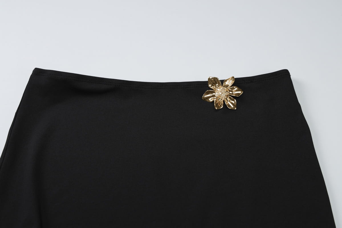 Black satin slim oblique shoulder mermaid dress featuring a gold-toned floral brooch with detailed petals and a sparkling center.