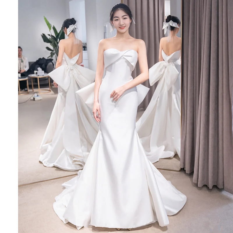 A sleek white satin mermaid wedding dress with a strapless bodice featuring a large bow at the neckline and a small trailing temperament flowing train.