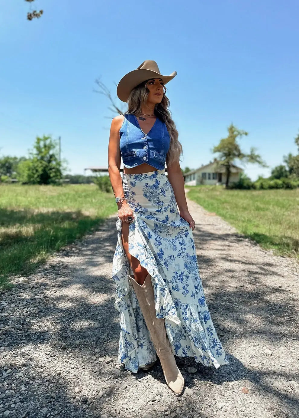 Woman wearing a white blue floral split print pleating big hem skirt paired with a blue denim crop top beige knee-high boots and a tan cowboy hat.