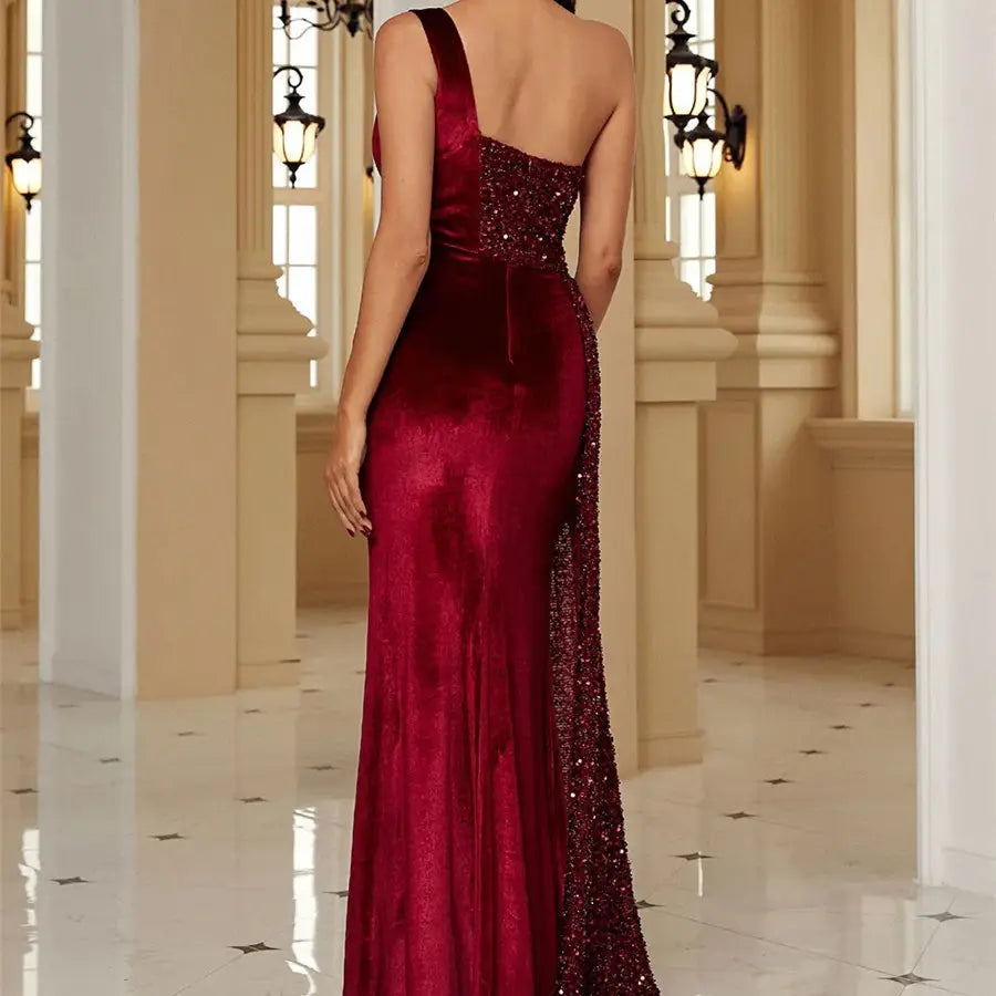 A luxurious burgundy velvet evening dress with a single strapless one-shoulder design featuring sparkling sequins along the back and side slit.