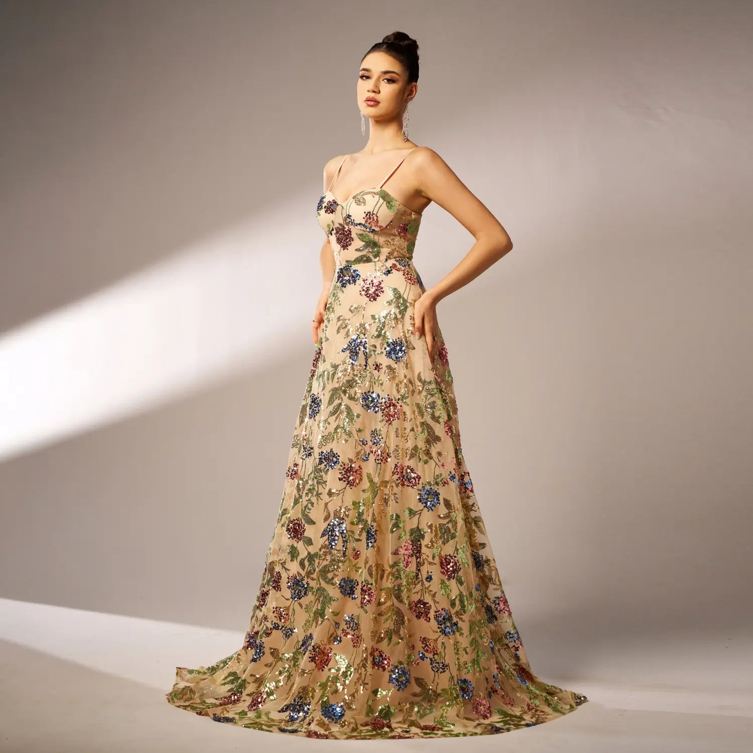 Elegant sleeveless sequined suspender evening dress in beige featuring a floral gown with sequined blossoms and green vines, thin spaghetti straps, and a flowing silhouette.