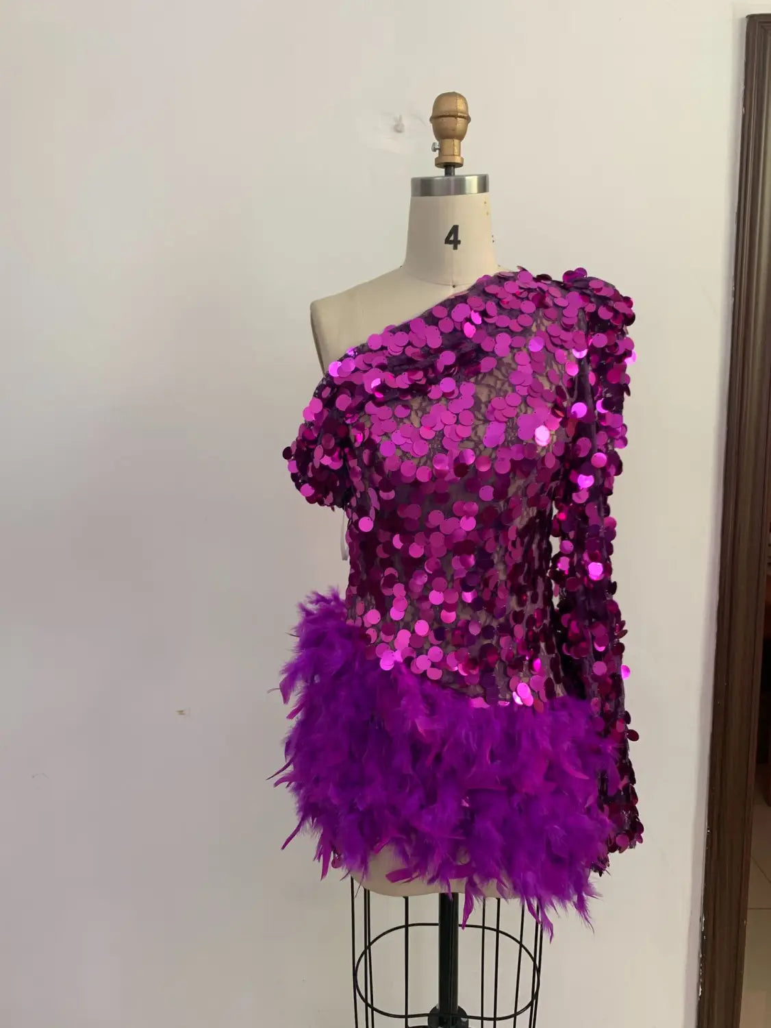 A vibrant purple sequined feather one-shoulder sleeve short dress displayed on a mannequin featuring a feathered hemline and sequined feather skirt detail.
