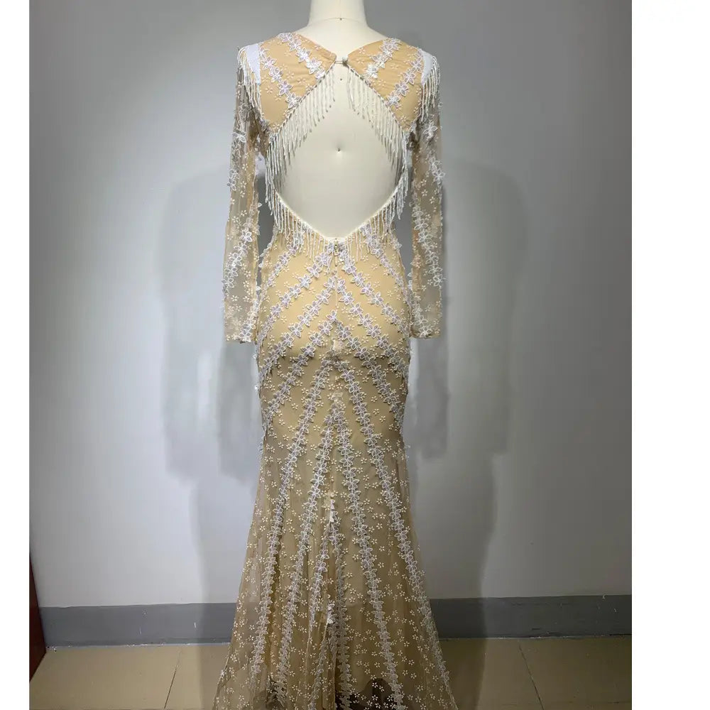 a shimmering champagne-colored backless tight fishtail gown featuring intricate white lace detailing and a dramatic open back with cascading fringe.