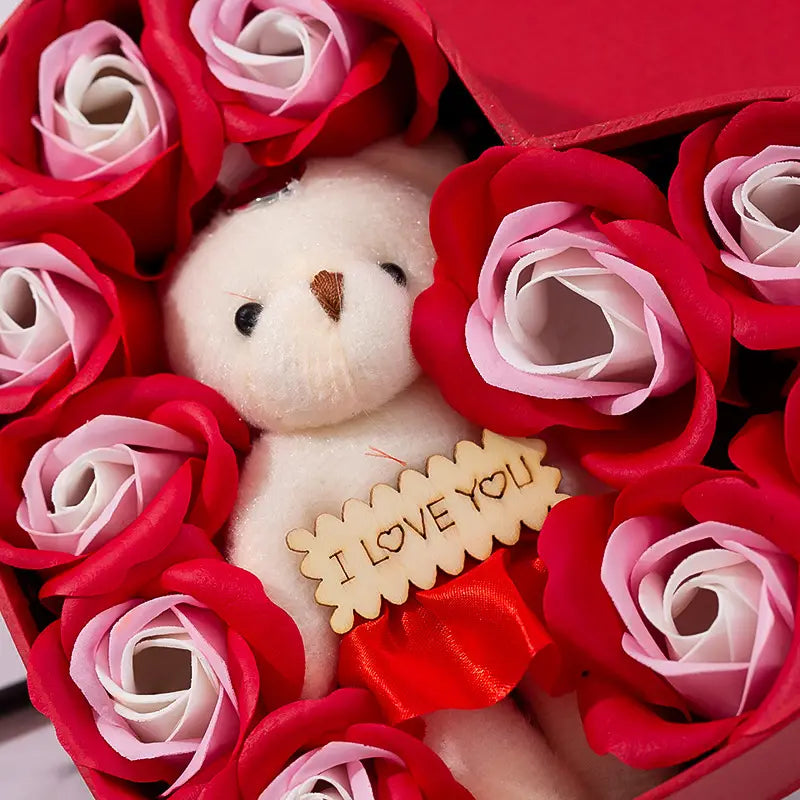 a plush white teddy bear with black eyes and a brown nose nestled among red and pink roses in a rose red heart gift box for mother’s day