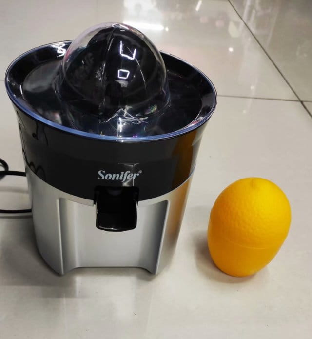 black and silver stainless steel electric lemon juicer with transparent dome top next to a bright yellow orange for efficient citrus juicing