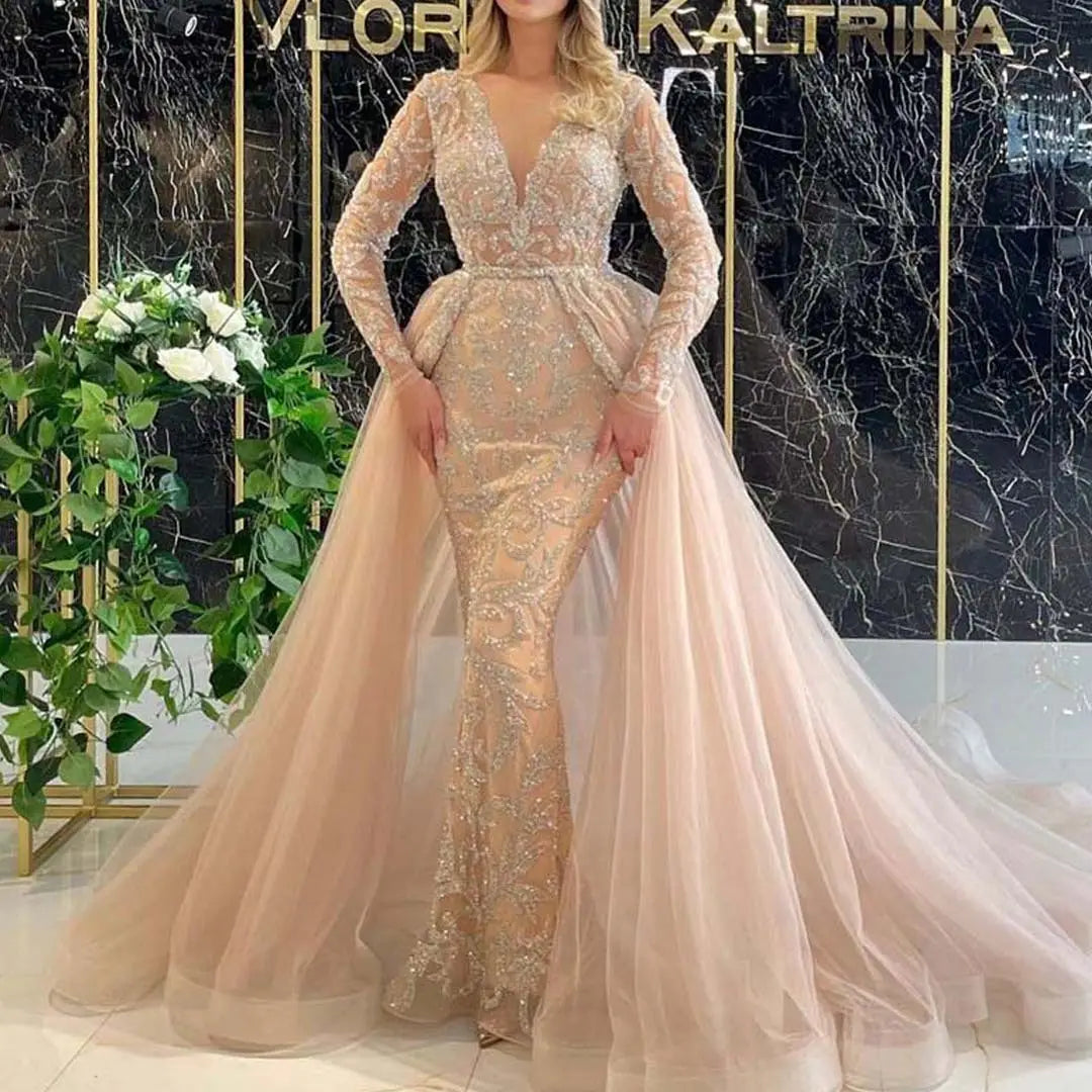 A blush pink mermaid-style wedding dress with long sheer lace sleeves, a beaded bodice, and an elegant flowing tulle train in a french style simple design.