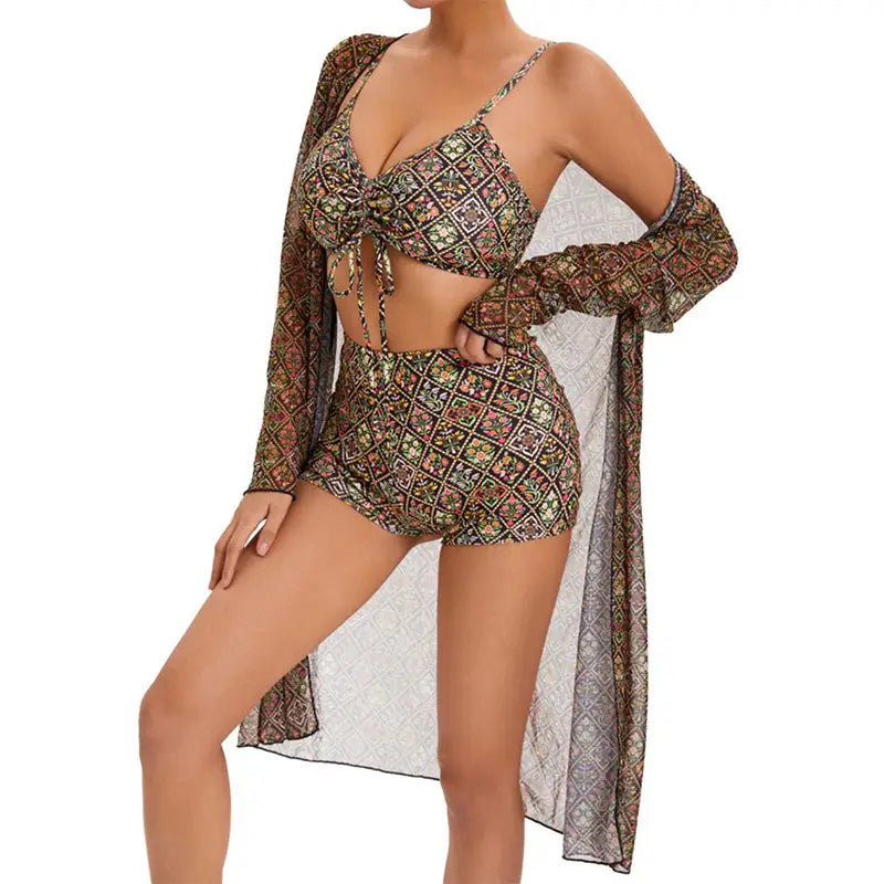 A woman wears a matching digital printing pleated tie bikini in earthy tones with intricate geometric patterns, paired with a sheer long-sleeved cover up featuring the same print.
