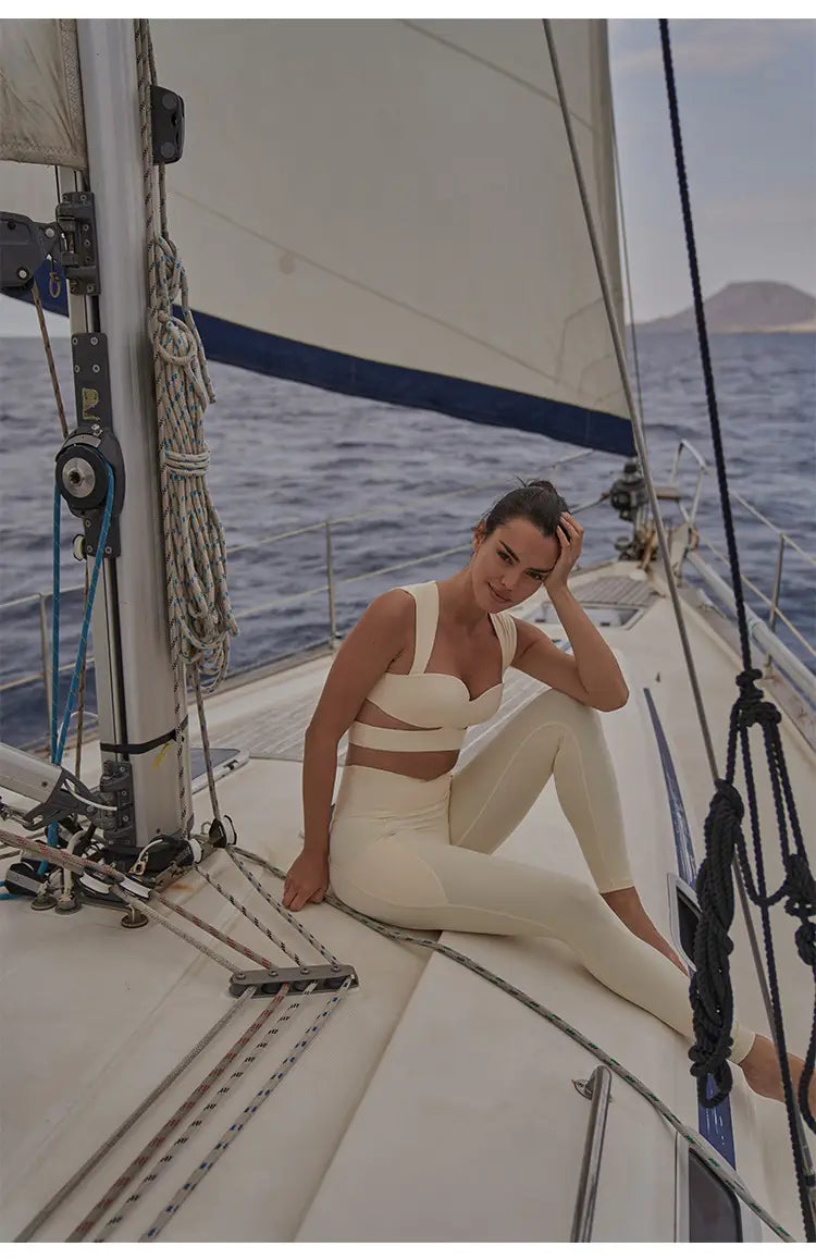 A woman wearing a cream-colored beige yoga pants and sport coat fitness set sits on the deck of a sailboat featuring the sport coat in a deep black color.