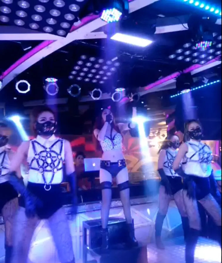 The central performer wears a white leather perform dance costume featuring a black-laced bodysuit with matching thigh-high stockings and dark boots, designed as a studded leather harness perform dance costume.