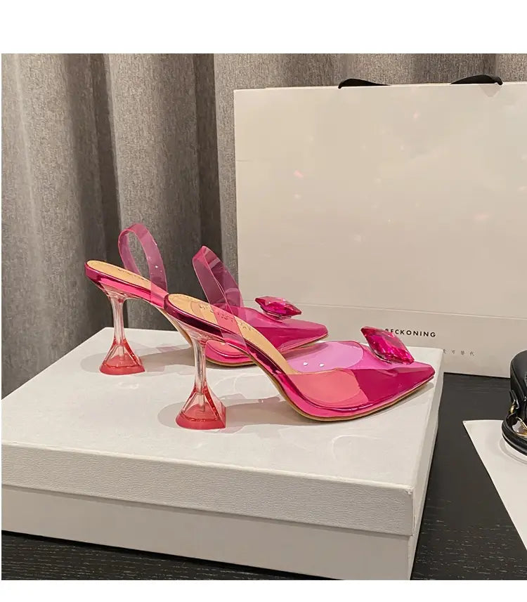 Vibrant pink satin bow women’s high-heeled sandal features transparent glossy material with crystal-like embellishments.