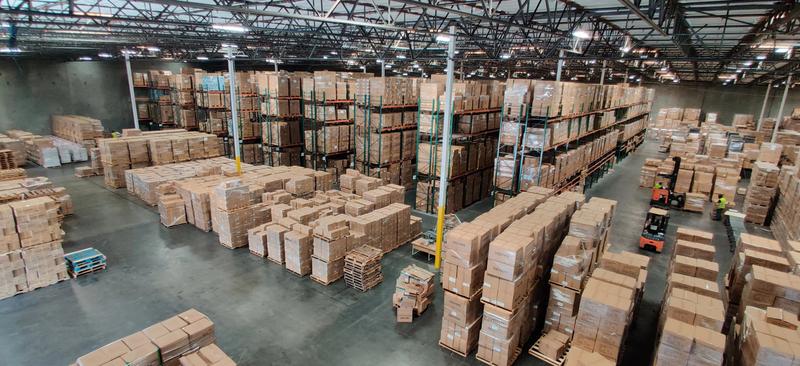 a large warehouse with metal shelving units holding stacked cardboard boxes and forklifts in aisles, highlighting storage of pink kiss stainless steel handle straw insulated tumblers designed to prevent stainless steel spill