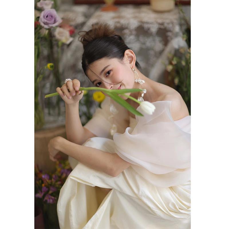A woman wears an ivory satin outdoor travel shooting gown with voluminous off-the-shoulder sleeves holding a single white tulip.