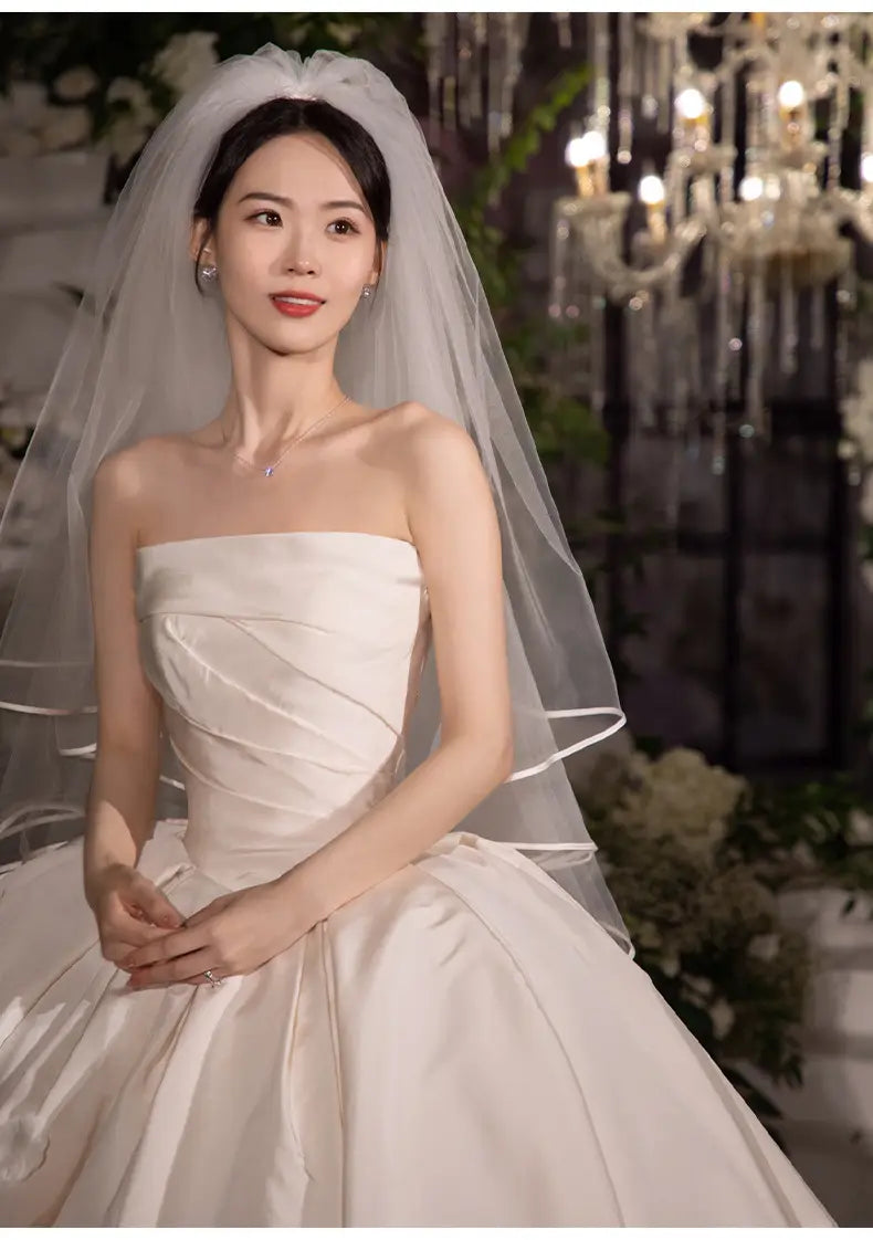 A simple elegant french style strapless white satin wedding dress with a fitted bodice, voluminous skirt, and complemented by an elegant sheer veil.