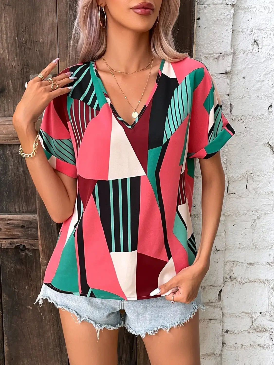A vibrant geometric-patterned women’s clothing printed v-neck top in shades of pink, green, black, and white with short sleeves.