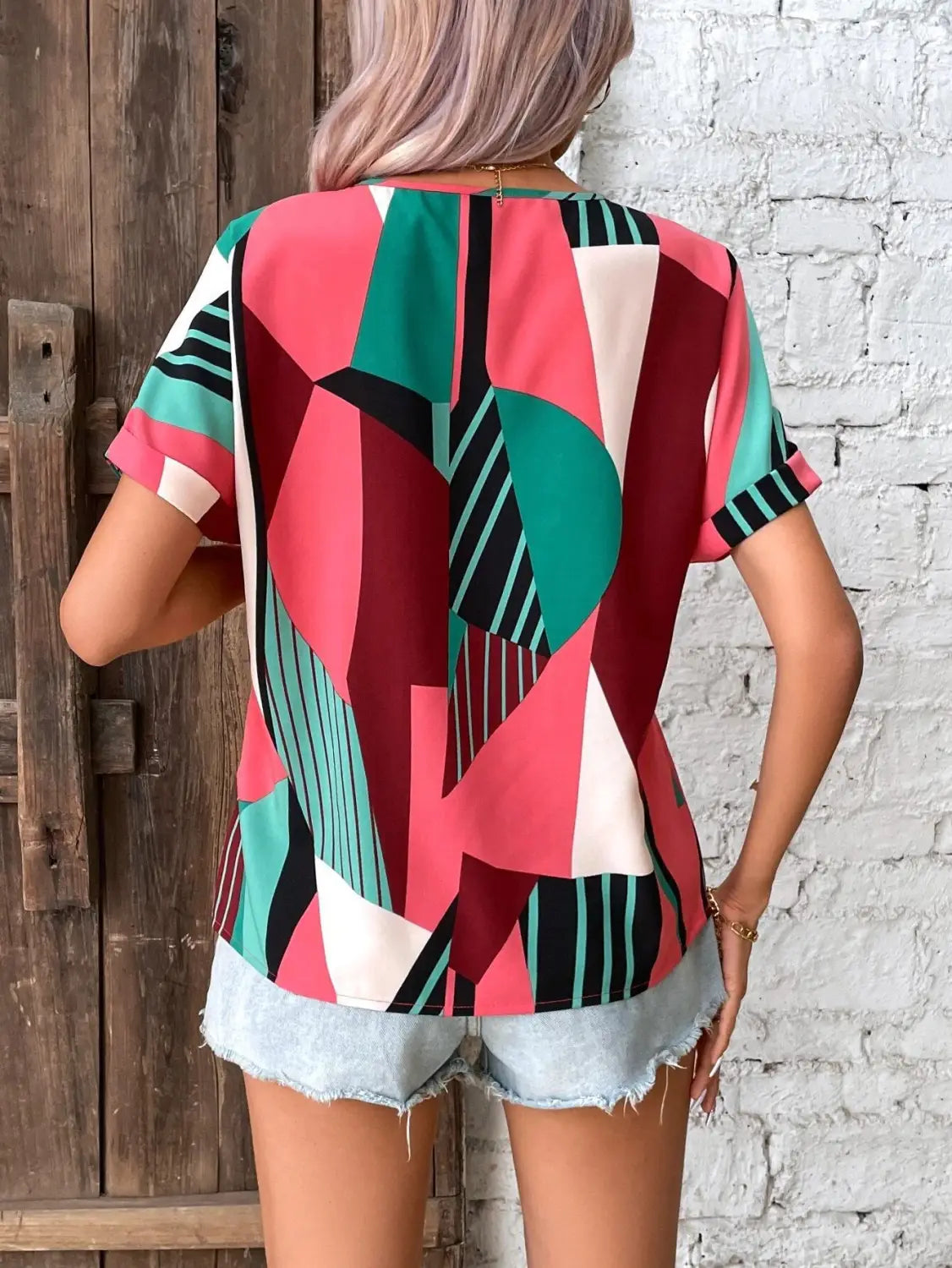 A vibrant geometric patterned women’s clothing printed v-neck top with short sleeves in shades of pink green maroon and black featuring a relaxed fit.