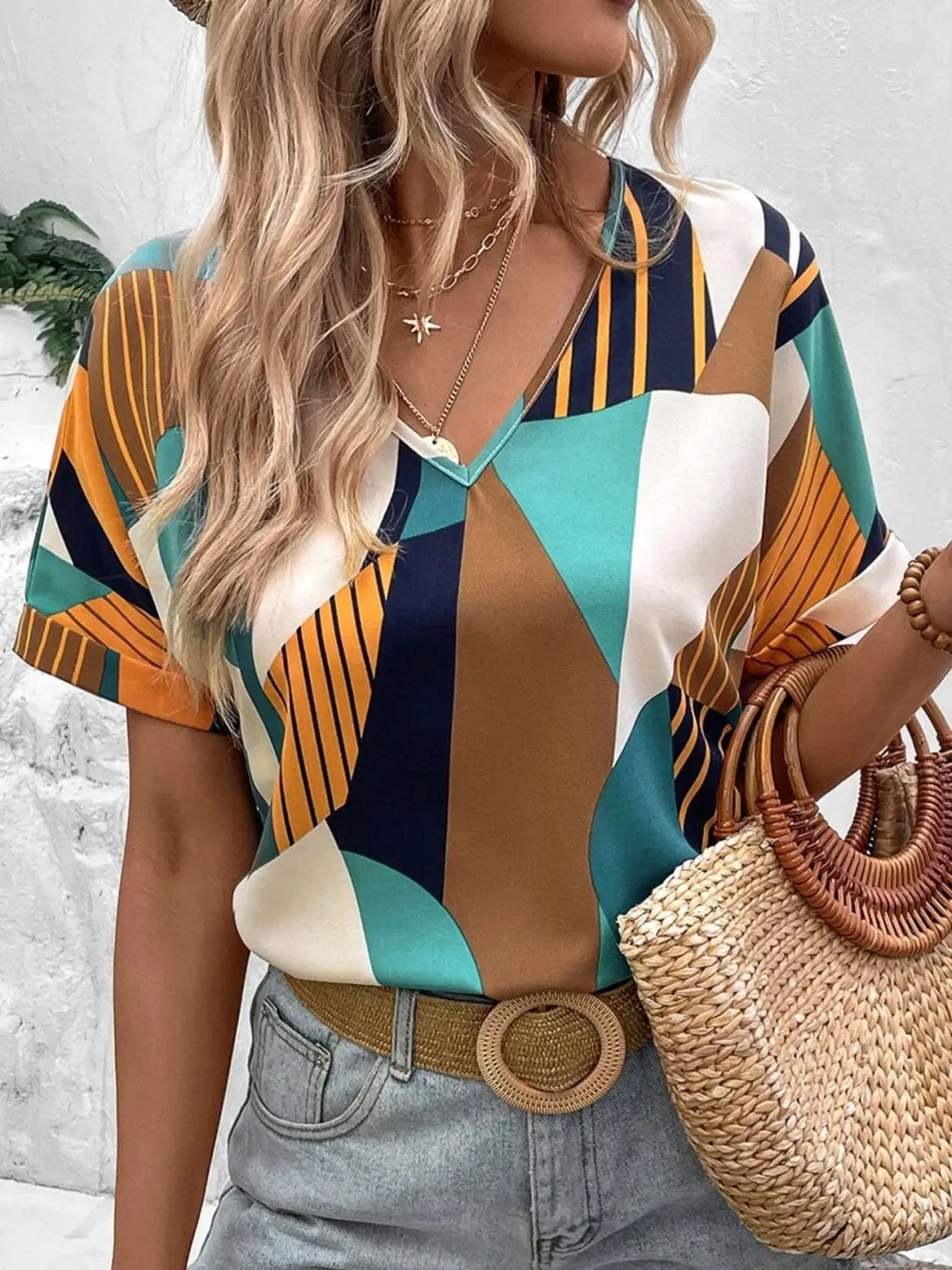 A woman wears a clothing printed v-neck short-sleeved blouse with bold geometric patterns in teal orange navy blue and cream from the geometric women’s clothing printed collection.
