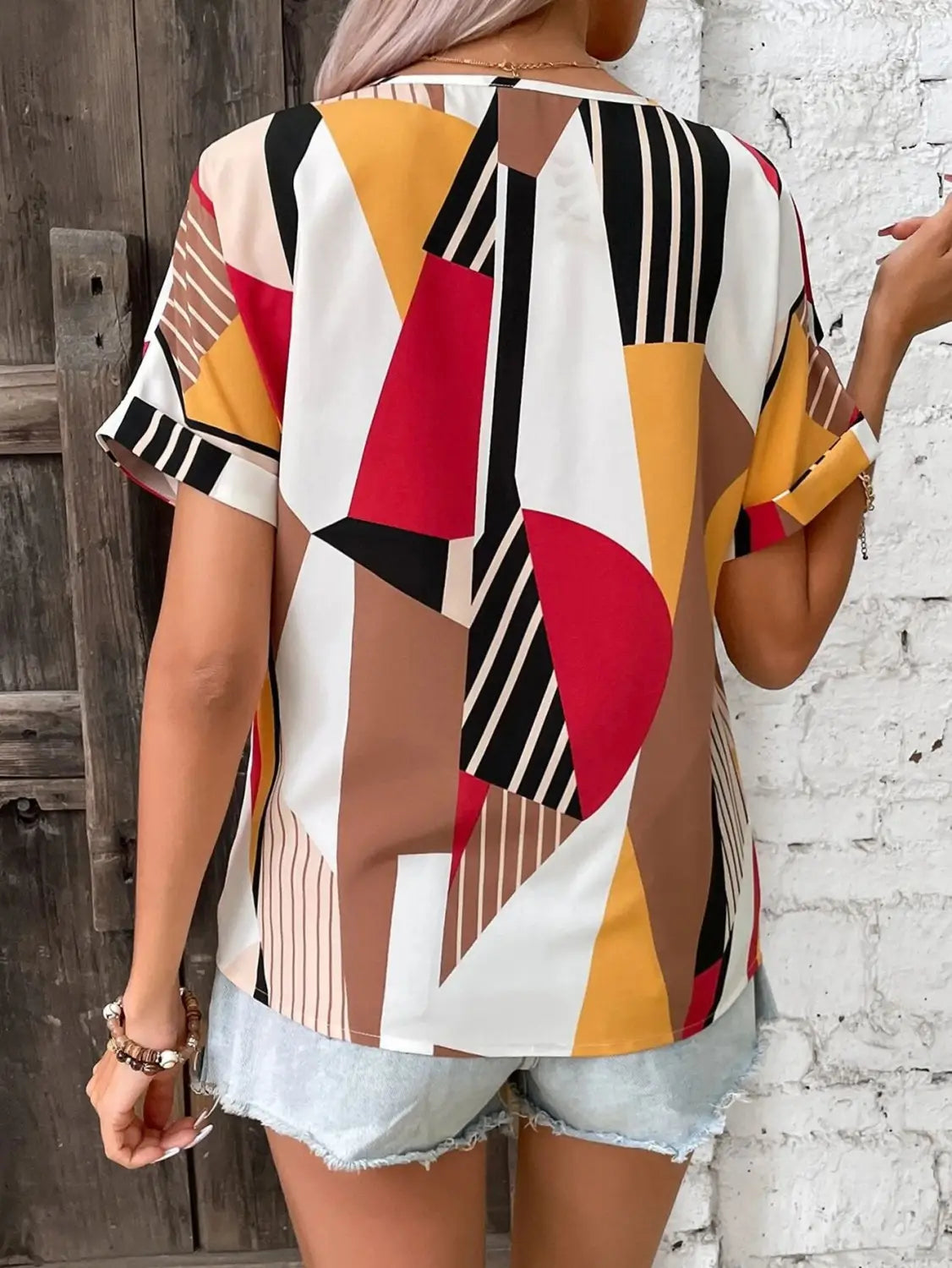 A vibrant geometric-patterned women’s clothing printed v-neck short-sleeve top in white, red, black, yellow, and beige with a relaxed fit.