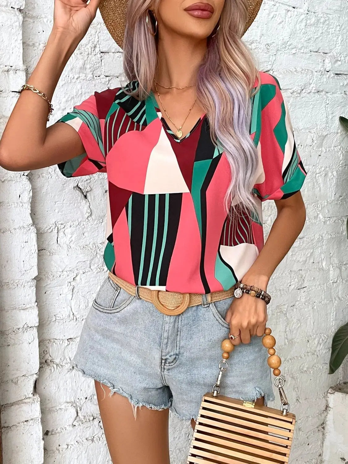 A vibrant geometric-patterned women’s clothing printed v-neck top featuring pink, green, black, and white colors with short sleeves.