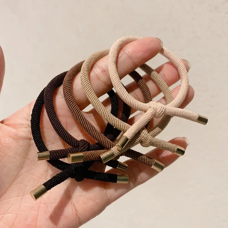 A hand holds a collection of beige brown and black high elasticity braided wrist hair ropes with metallic gold caps in a 5 pack.