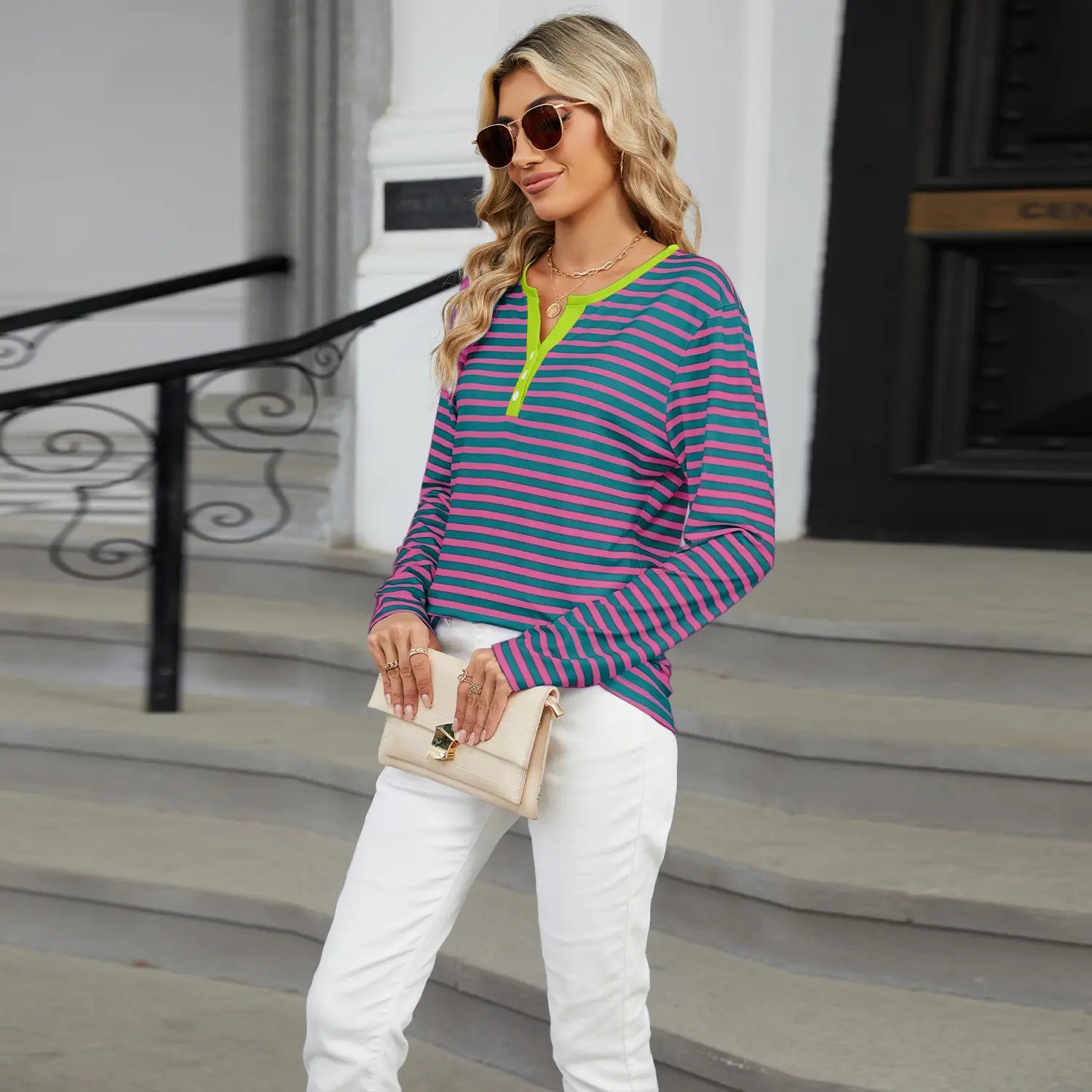 A woman wearing a women’s v-neck striped loose henley top with pink and teal horizontal stripes and a neon green v-neck collar.